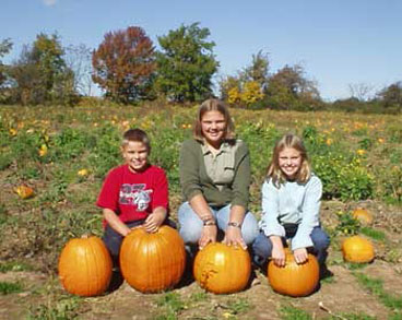 Three kids sitting on big orange pumpkins in the patch